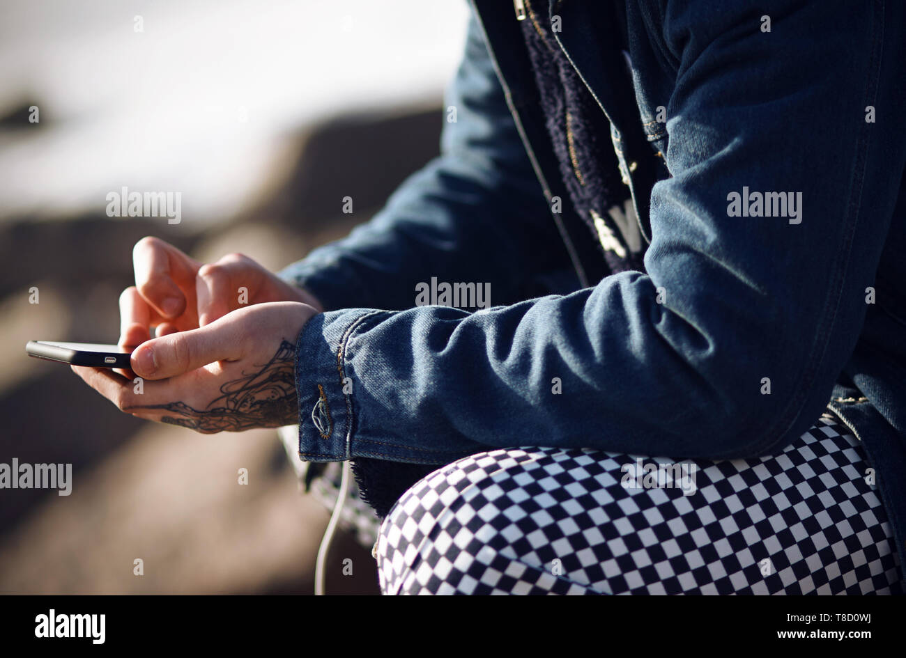 A fashionably dressed man in a denim jacket and plaid pants holds a touch phone in his tattooed hands Stock Photo