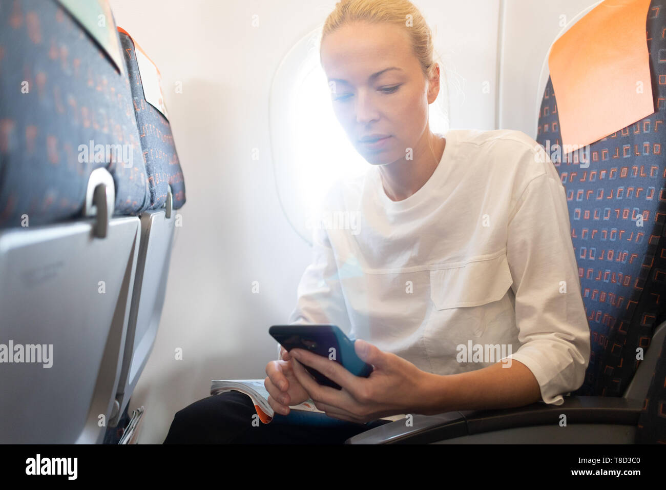 Young woman using mobile phone on airplane. Female traveler reading on her phone on the aircraft seat near the window during the flight in the Stock Photo