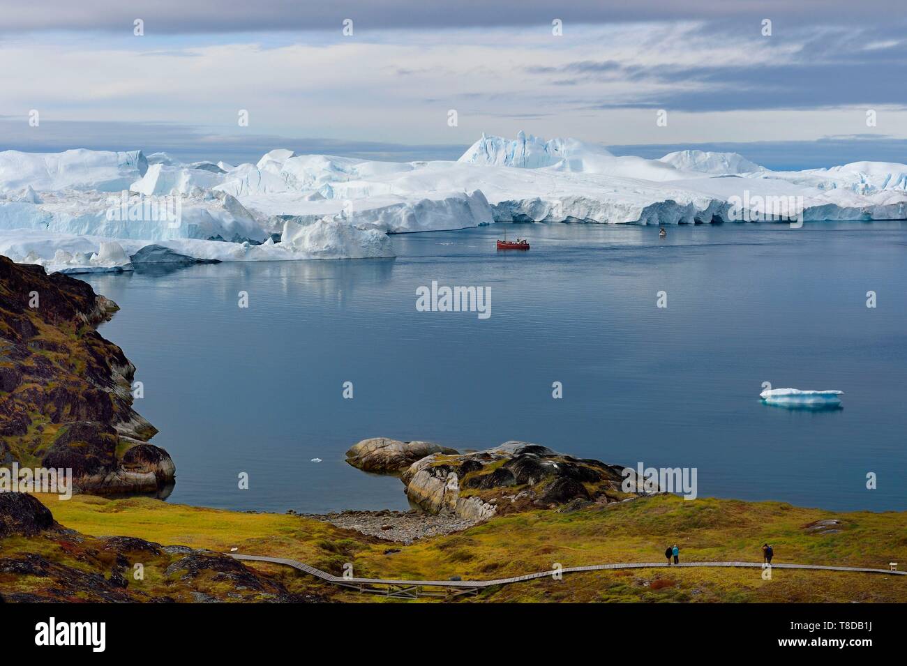 Greenland, west coast, Disko Bay, Ilulissat, icefjord listed as World heritage by UNESCO that is the mouth of the Sermeq Kujalleq Glacier (Jakobshavn Glacier), hiking on the wooden walkway going to the Sermermiut site and fishing boat at the foot of icebergs Stock Photo