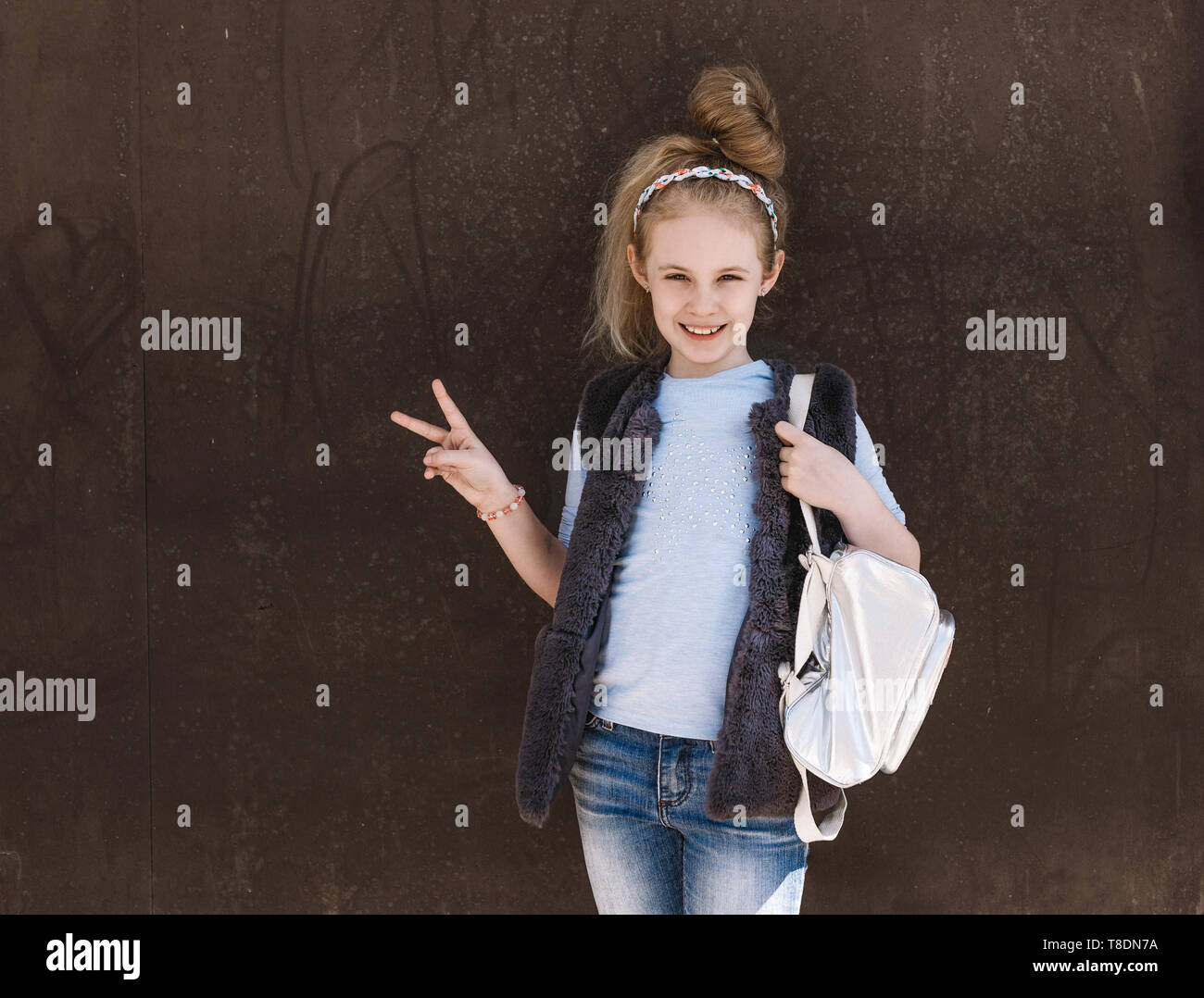 Charming eight-year-old girl in a trendy outfit with a backpack standing on the street on a sunny day. Stock Photo