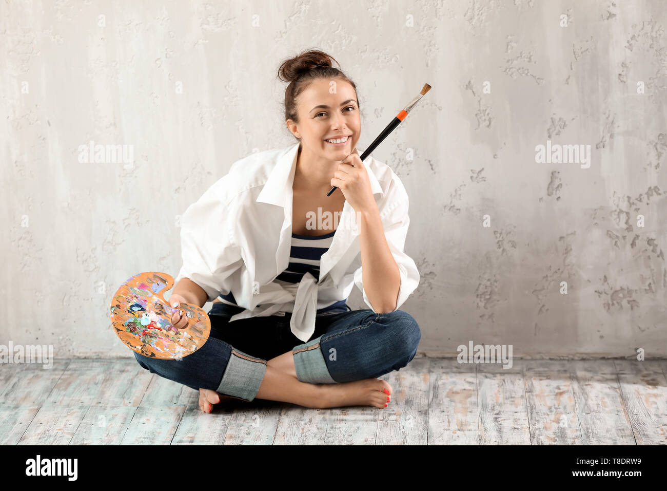 Female artist with brush and paint palette sitting on floor near grey ...