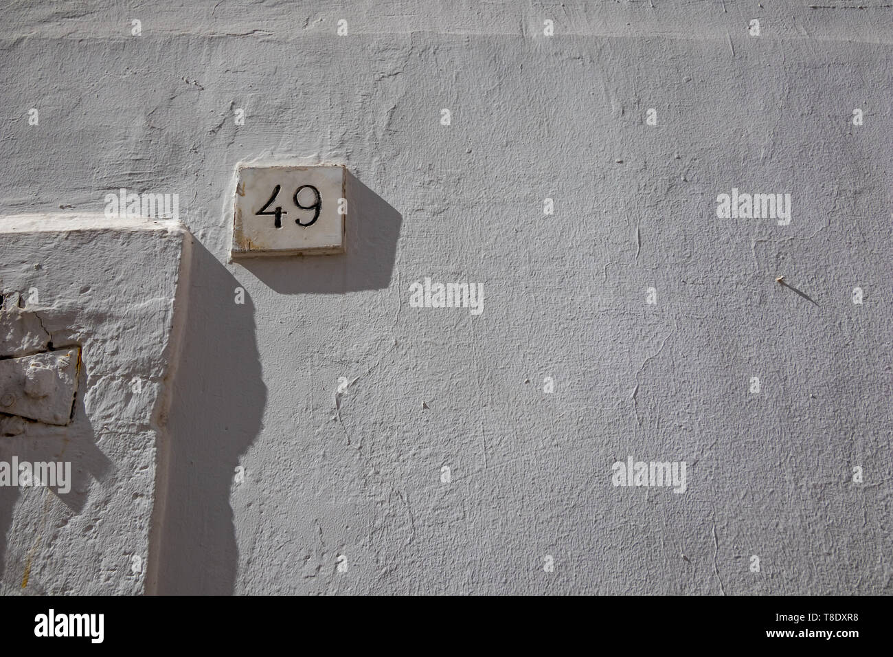 White house street number forty-nine 49 carved into marble parallelepiped full with black paint, white-washed buildings in Noci, Puglia, Italy with summer day shadows Stock Photo