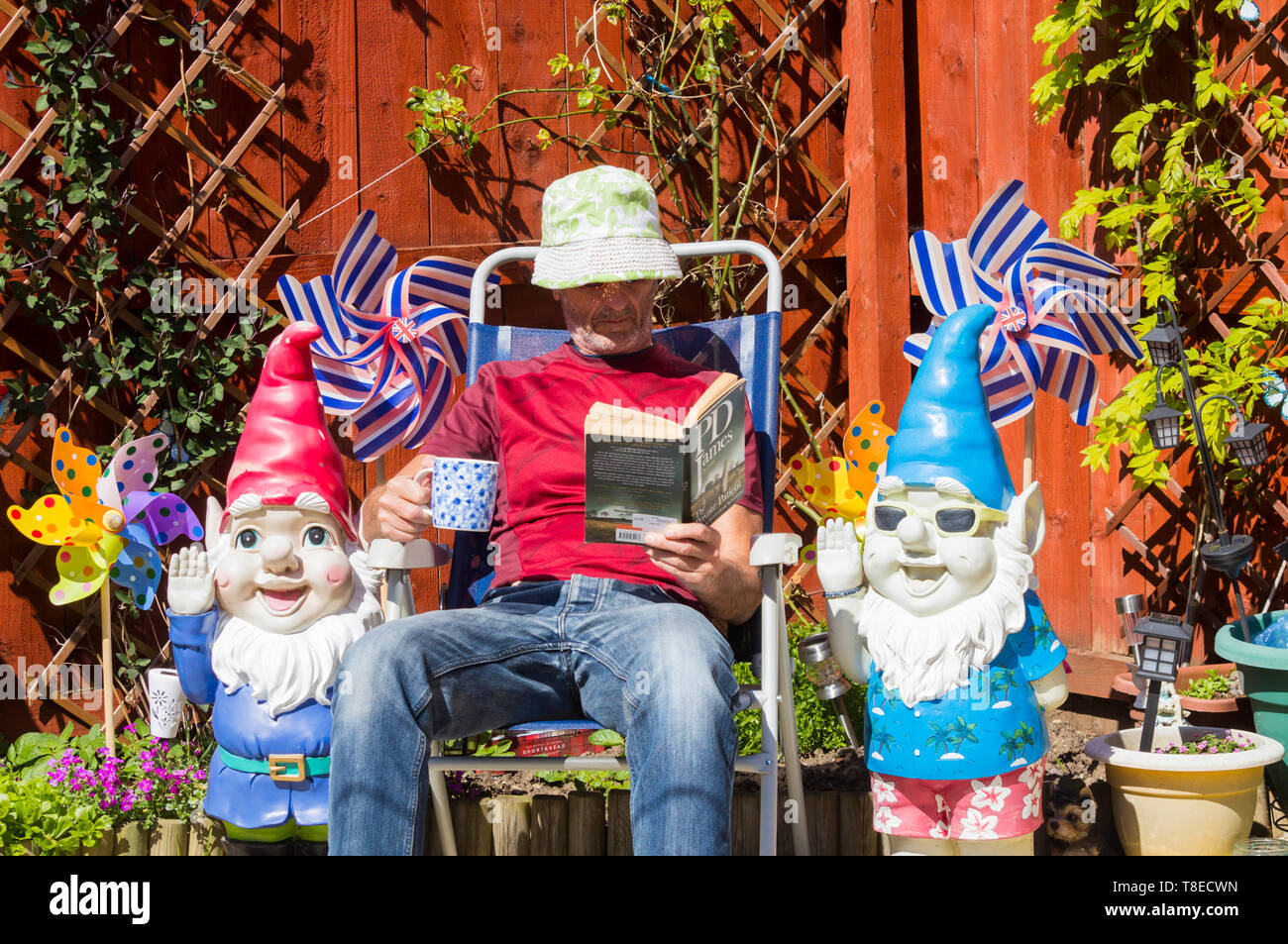 Mature man relaxing in garden flanked by garden gnomes. Summer, heatwave. UK Stock Photo