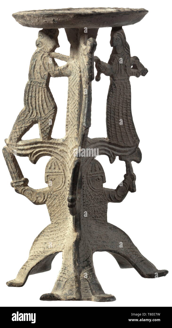 A North European Romanesque candle holder, 11th/12th century Pricket candle holder on three feet made from a heavily leaded pewter alloy. The base made of three sitting warrior figures with Norman/Anglo-Saxon full-length mail shirts with nasal helmet and aventail. Two of the figures with raised sword, the third one with an axe. Above that a group of figures, a woman leading a pinioned captive, holding a bird in her right hand. The third figure is a man with sword, blowing a hunting horn. Slightly curved drip pan with blossom-shaped decoration and, Additional-Rights-Clearance-Info-Not-Available Stock Photo