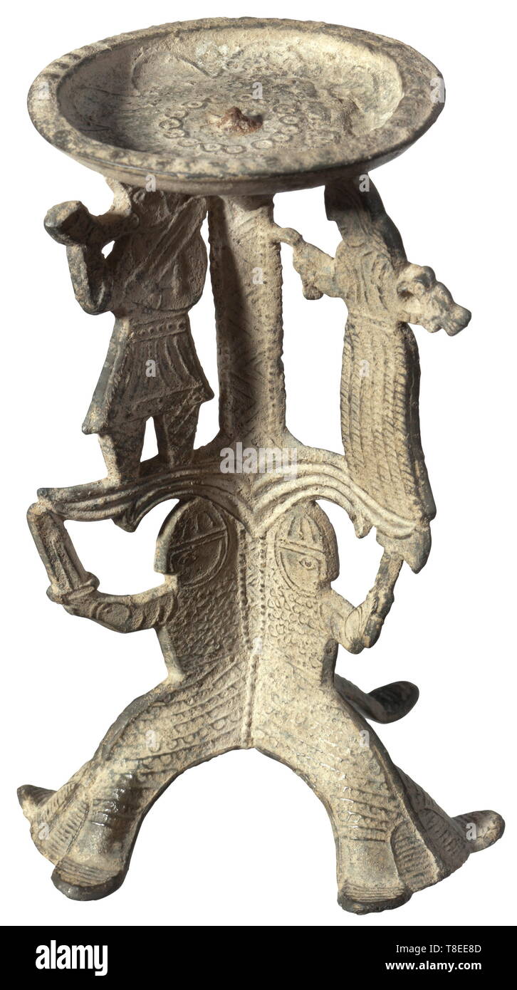A North European Romanesque candle holder, 11th/12th century Pricket candle holder on three feet made from a heavily leaded pewter alloy. The base made of three sitting warrior figures with Norman/Anglo-Saxon full-length mail shirts with nasal helmet and aventail. Two of the figures with raised sword, the third one with an axe. Above that a group of figures, a woman leading a pinioned captive, holding a bird in her right hand. The third figure is a man with sword, blowing a hunting horn. Slightly curved drip pan with blossom-shaped decoration and, Additional-Rights-Clearance-Info-Not-Available Stock Photo