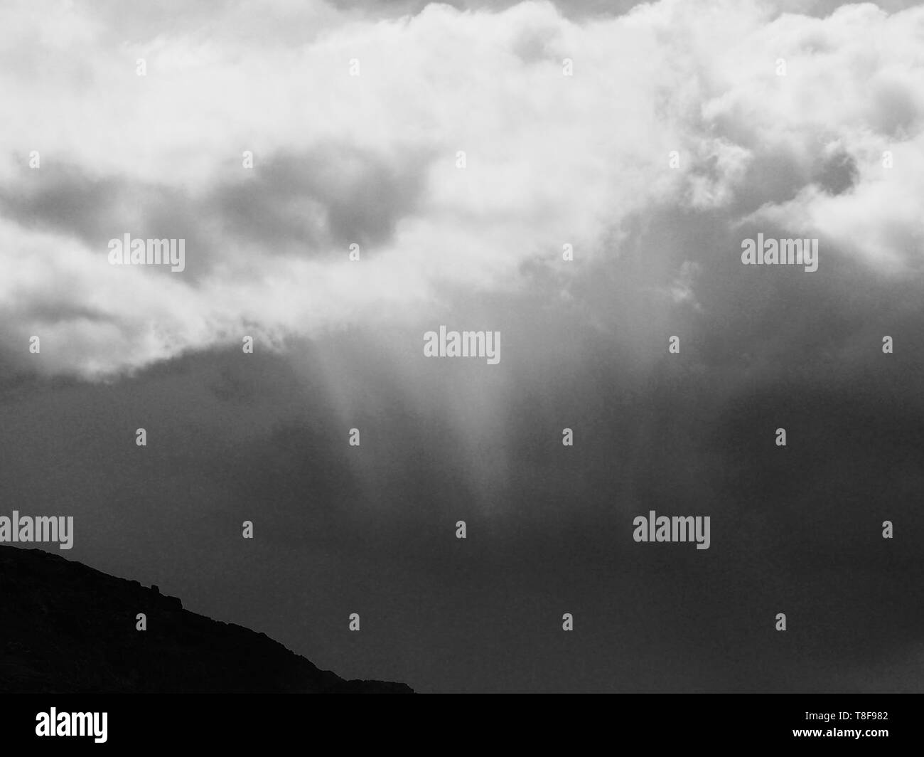 Shades of grey, Wispy clouds and beams of sun light streaming and breaking through the fluffy cumulus clouds above pointing towards the ground Stock Photo