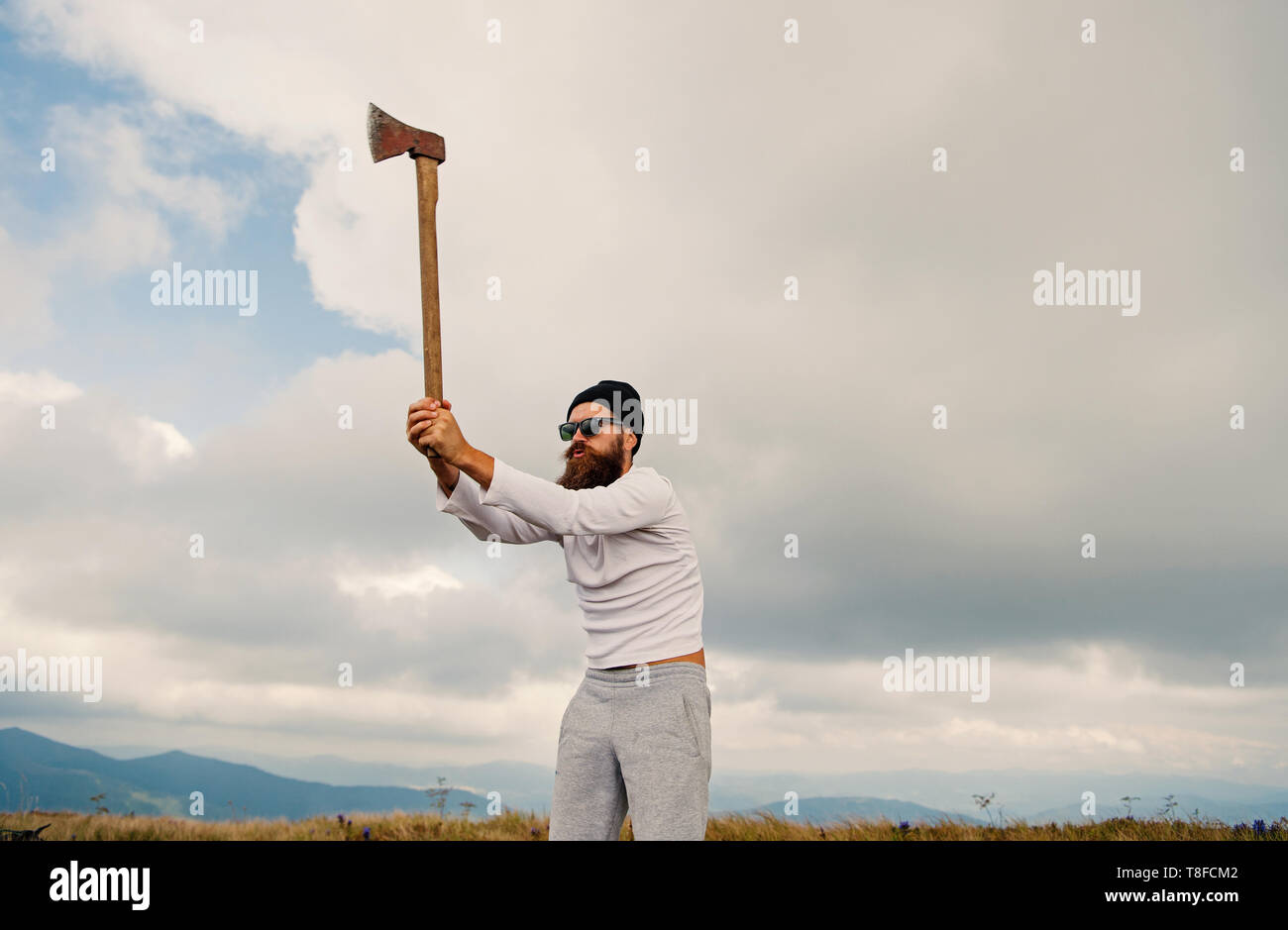 Man bearded lumberjack in sunglasses, hat raise axe on mountain landscape on cloudy sky. Logging and chopping concept. Wanderlust, hiking, vacation, travelling. Stock Photo