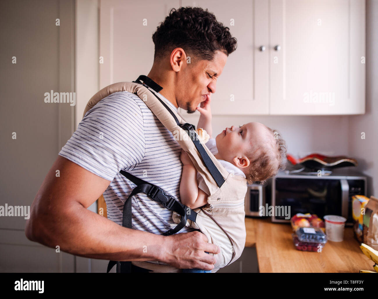 A father with small toddler son in carrier in kitchen indoors at home. Stock Photo