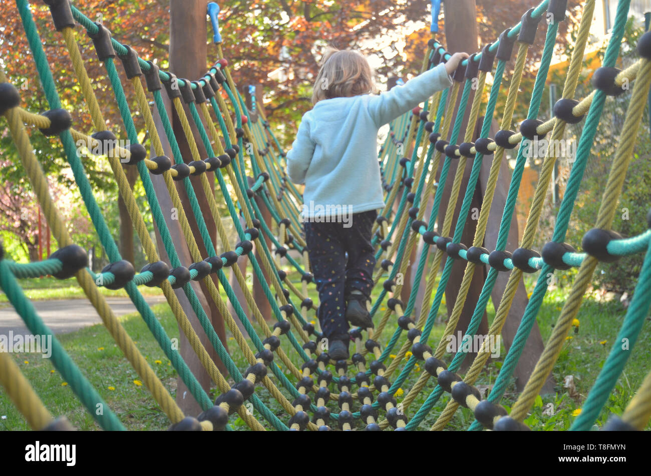 Cute little cca 3 year old girl walking on a net and rope  bridge-like play structure, on a sunny spring playground afternoon, back view. Stock Photo