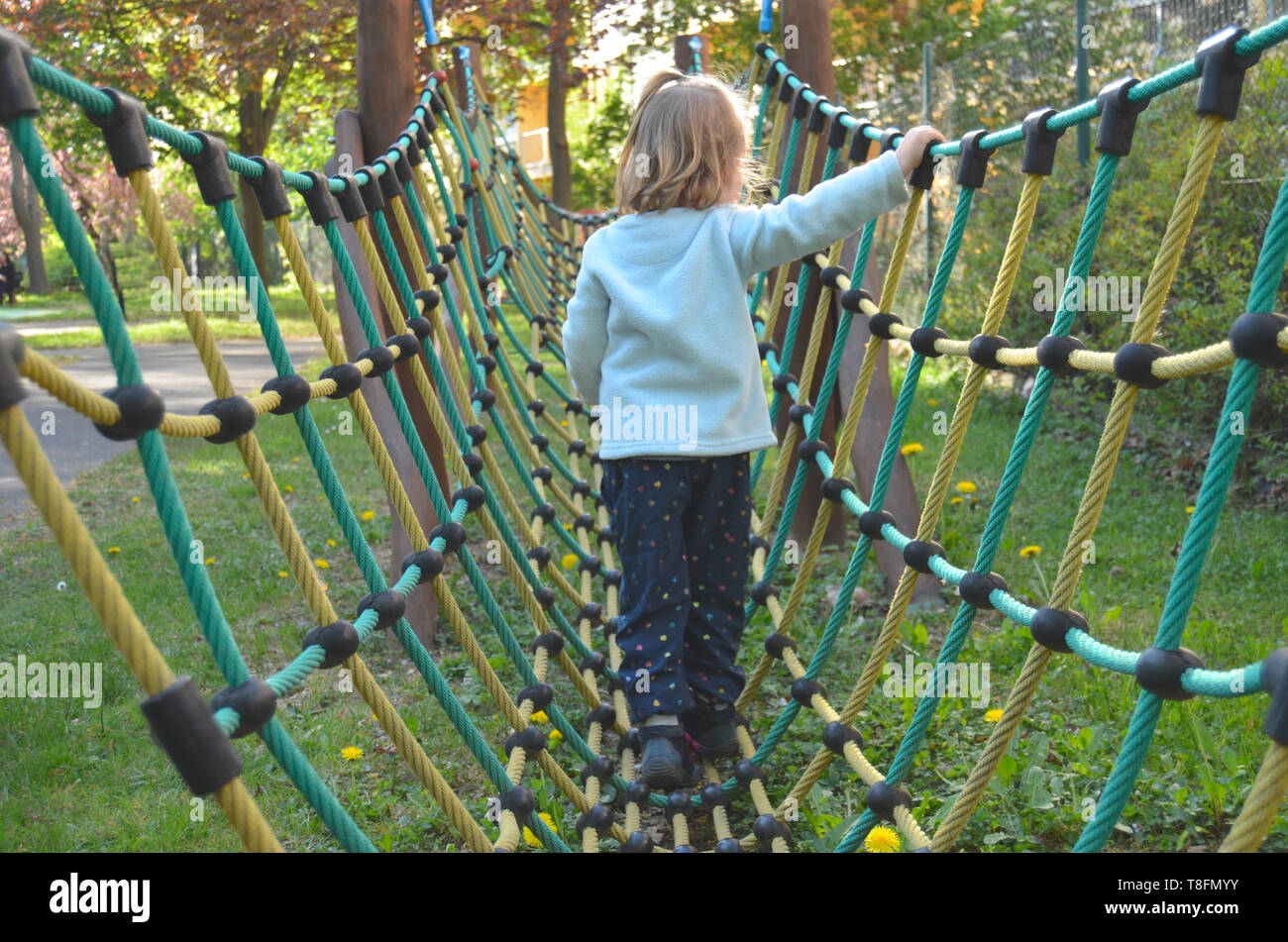 Cute little cca 3 year old girl walking on a net and rope  bridge-like play structure, on a sunny spring playground afternoon, back view. Stock Photo