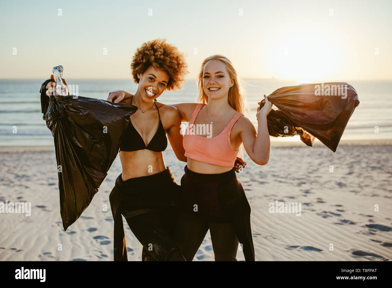 Two young female volunteers with garbage bag. Two surfers standing on the beach with plastic garbage bags with sunset in background. Stock Photo