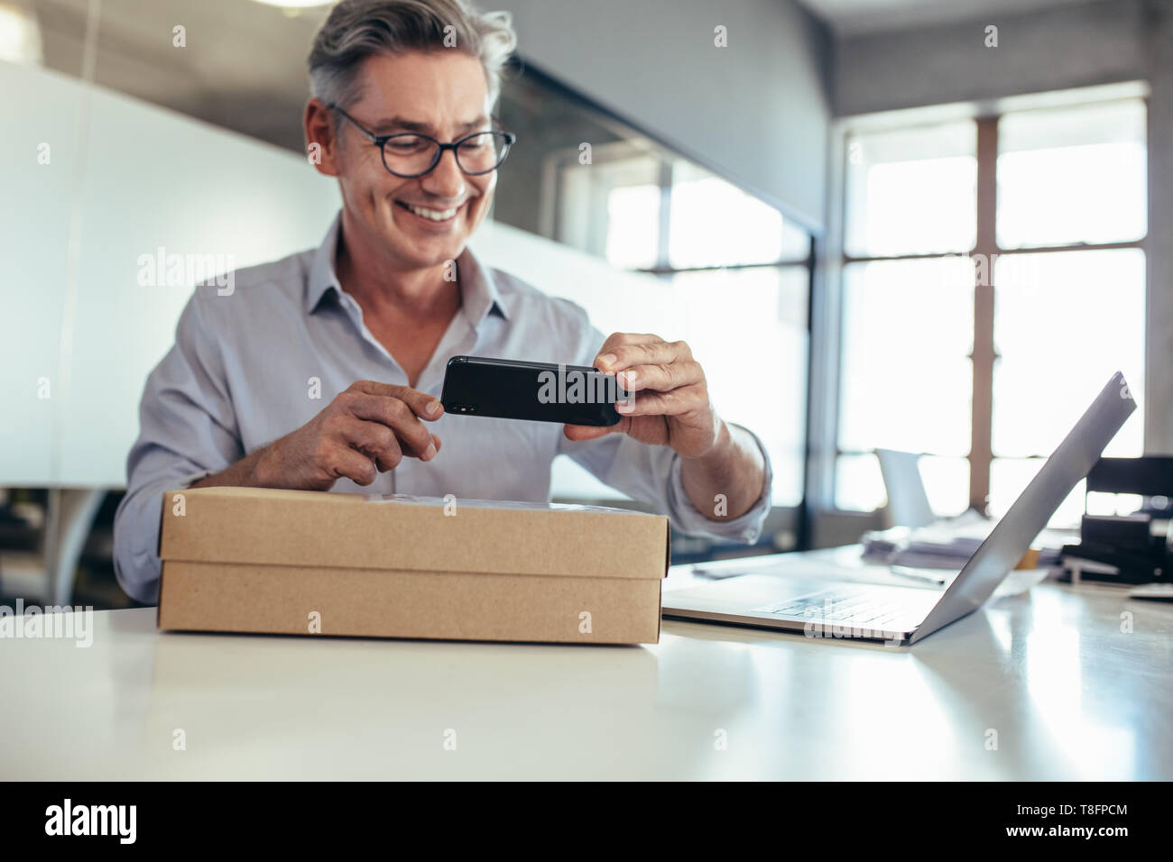 Smiling mid adult man taking scanning a delivery box on his desk. Man taking picture of a package. Stock Photo
