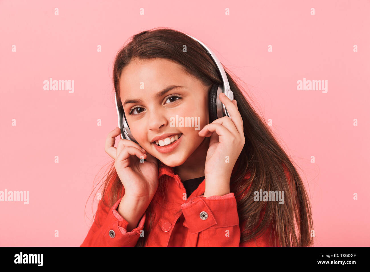 Image of caucasian little girl in casual wearing headphones listening to music isolated over red background Stock Photo