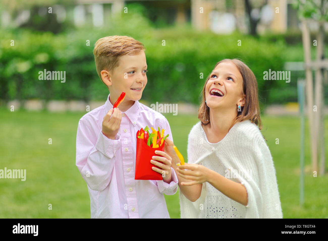 Happy kids eating fresh vegetables in nature - smiling children sharing colorful bio peppers sliced in form of french fries - little boy and girl Stock Photo