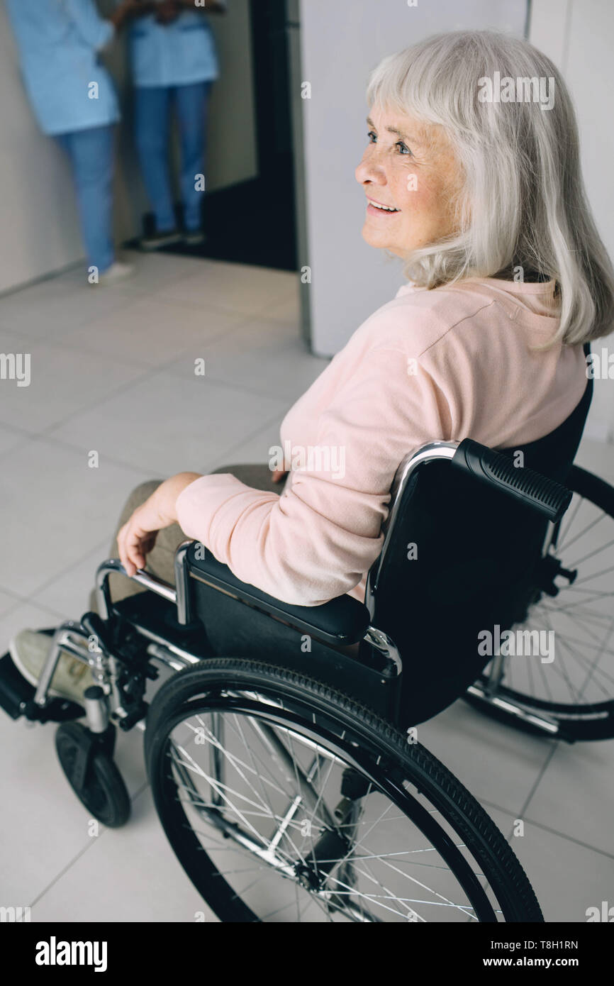 Smiling senior woman in wheelchair sitting at the hospital corridor. Go home after discharge from hospital Stock Photo