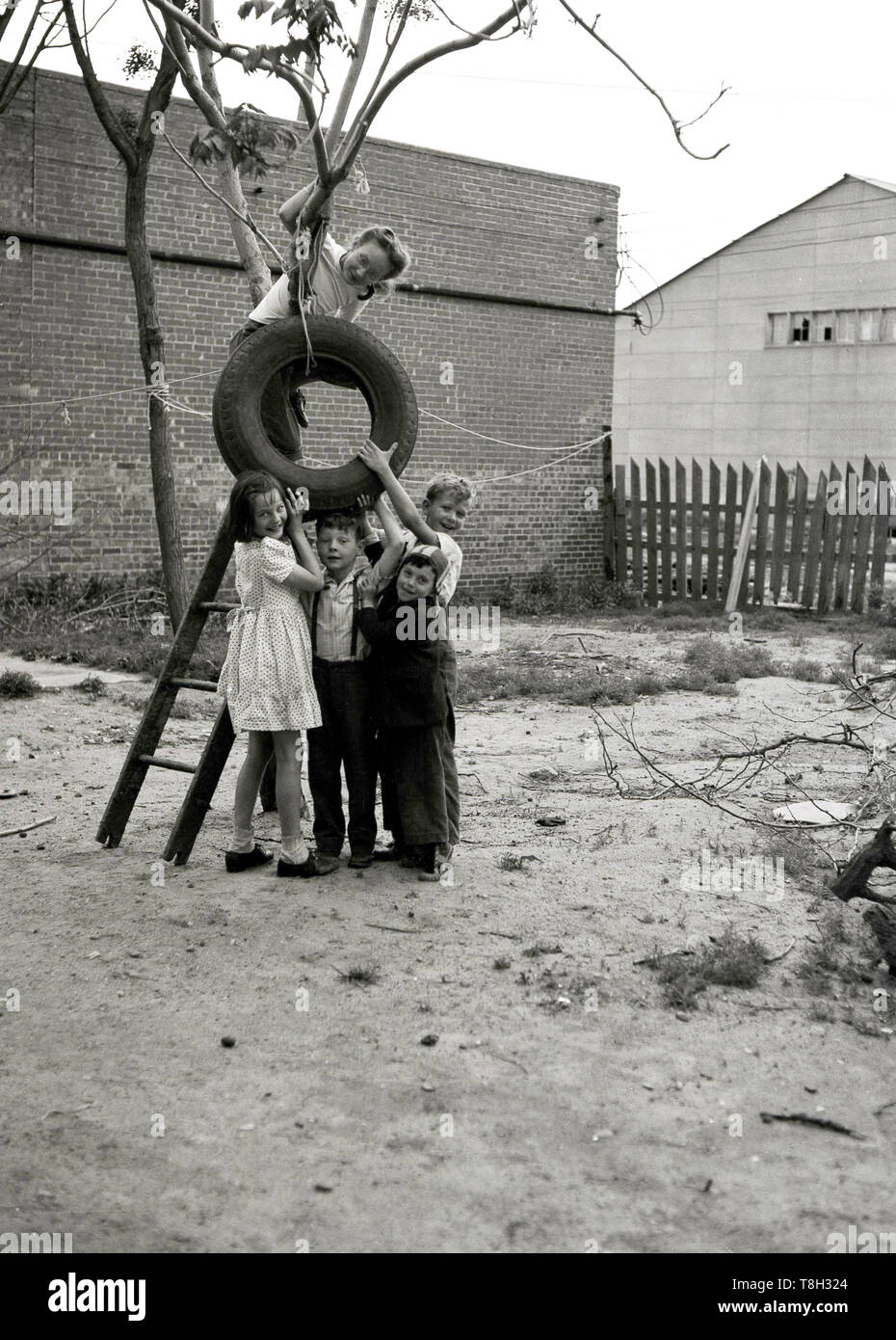 1970s, historical, hanging up a tyre swing....four young children standing together outside in a sandy industrial area of land or yard, holding up a rubber car tyre, while a young girl having climbed a ladder to get up a small tree, is trying to attach it to it with string.  Trye swings are great fun for to children to play on when growing up and it is also recycling. Stock Photo