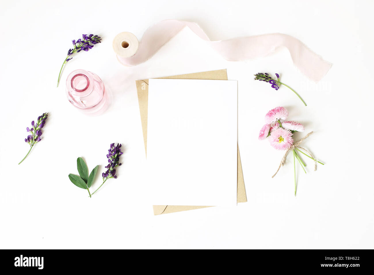 Feminine stationery, desktop mock-up scene. Vertical blank greeting card, craft paper envelope,silk ribbon, daisy and alfalfa flowers.White table Stock Photo
