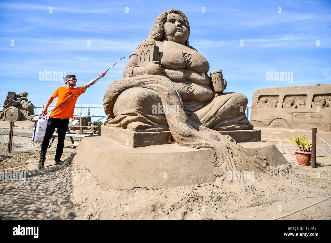 A worker sprays water to stop the sand drying out on a sculpture of an obese mermaid holding McDonald's fast food items, at the Weston Sand Sculpture Festival, Weston-super-Mare. Stock Photo