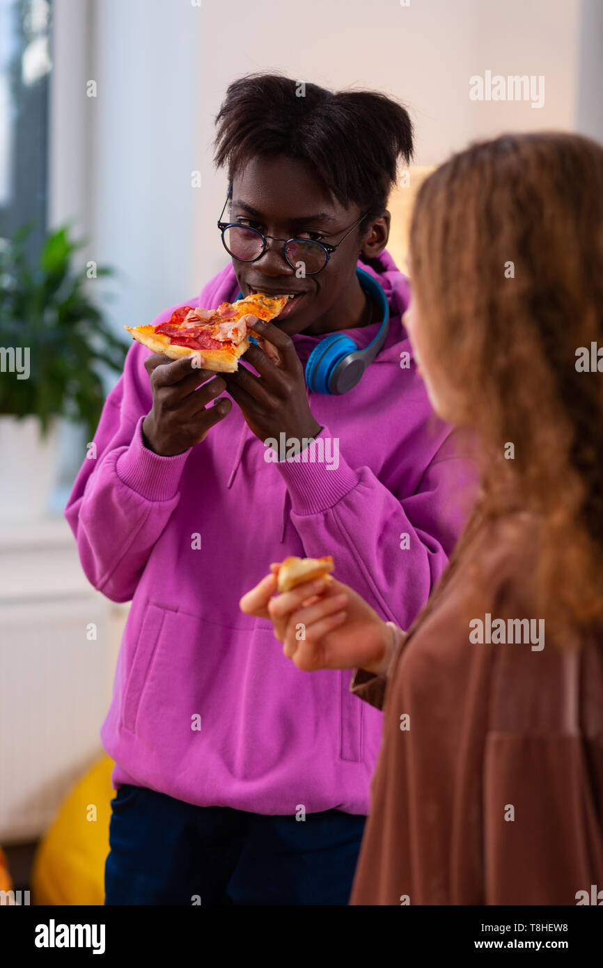 Teenager biting piece of pizza while eating with friend Stock Photo