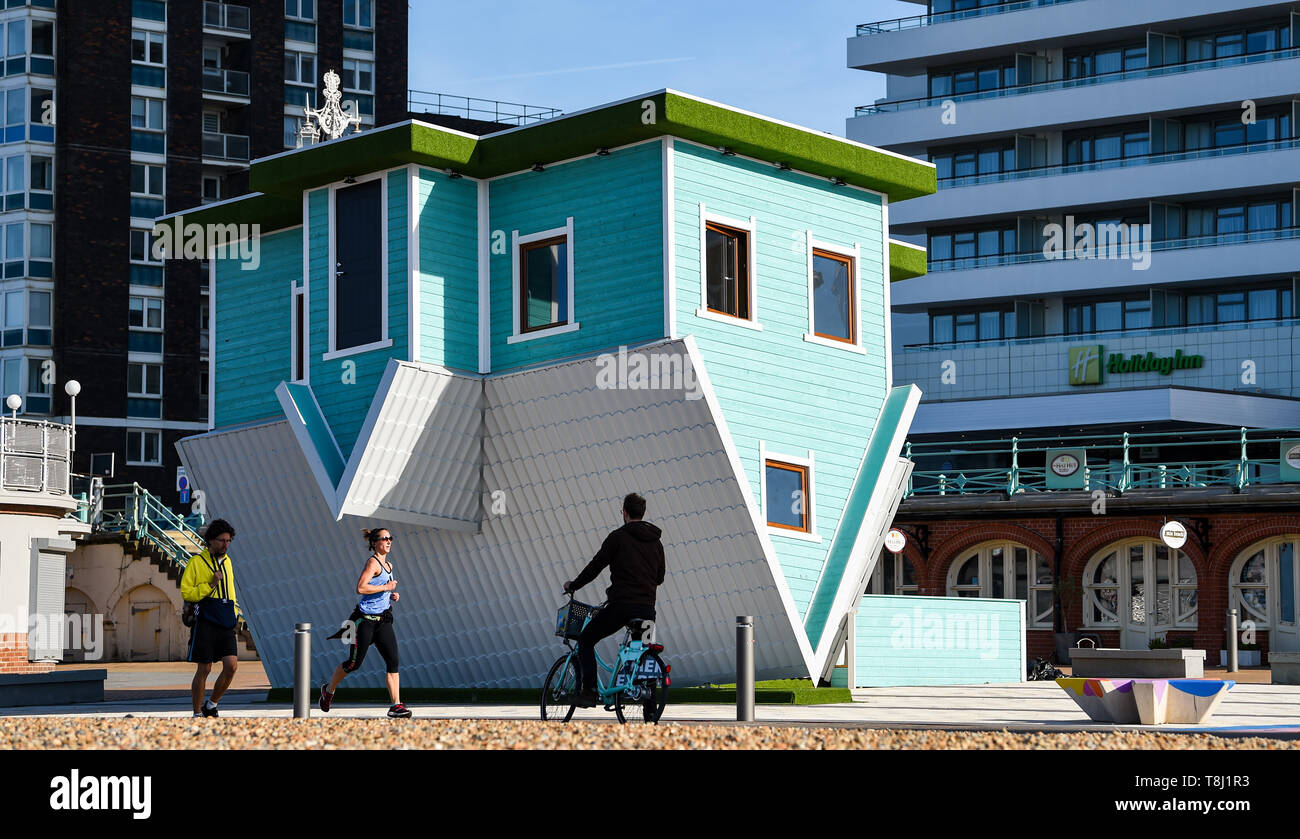 Brighton, UK. 14th May, 2019. Early morning runners and walkers by Brighton's newest tourist attraction the Upside Down House on the seafront near the West Pier on a beautiful sunny morning with temperatures reaching into the twenties in some parts of Britain today. The Upside Down House is due to open to the public this coming weekend . Credit: Simon Dack/Alamy Live News Stock Photo