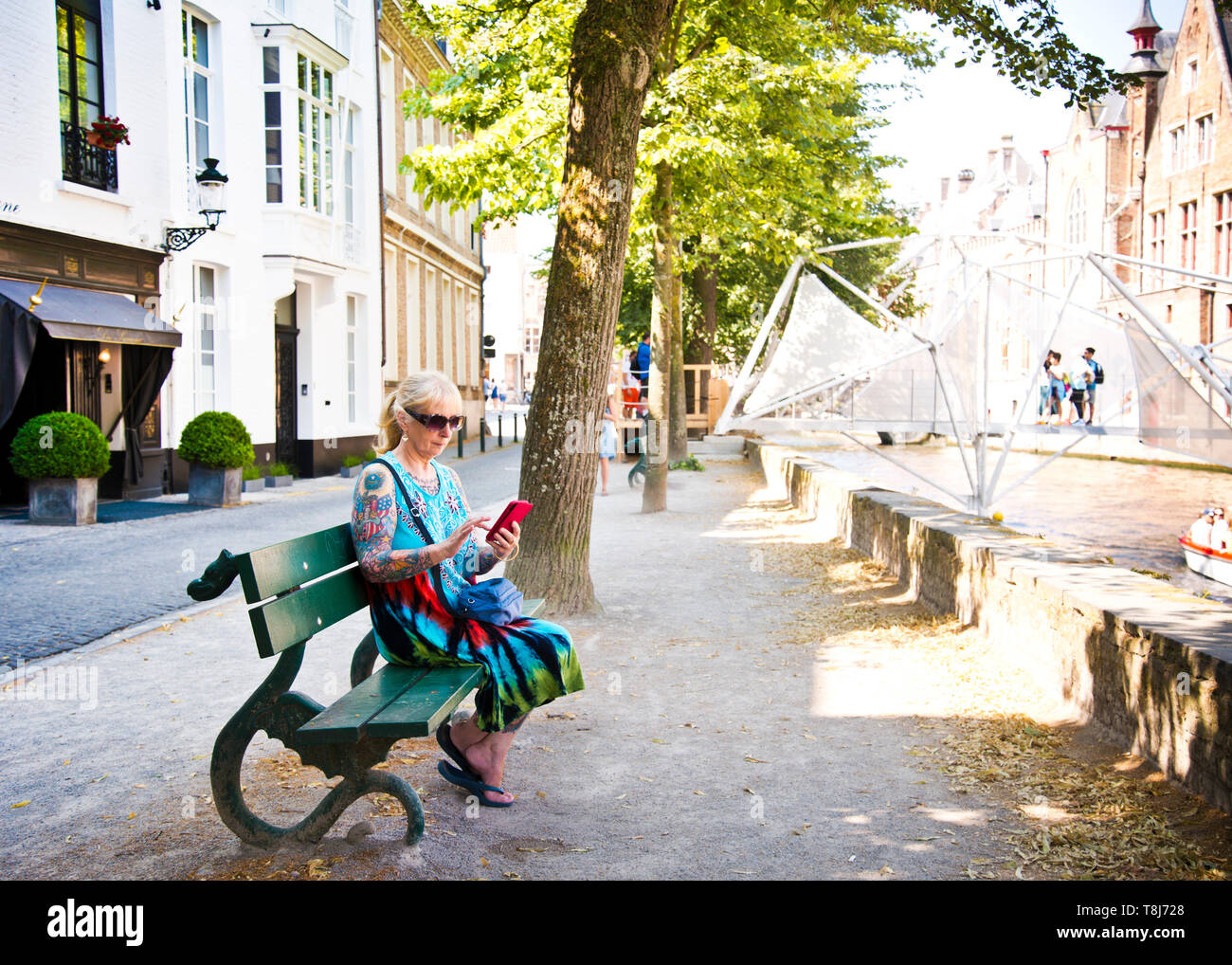 woman sitting alone on green public bench at canal bank,preparing to text  on mobile phone. city of Brugge, Belgium. Stock Photo