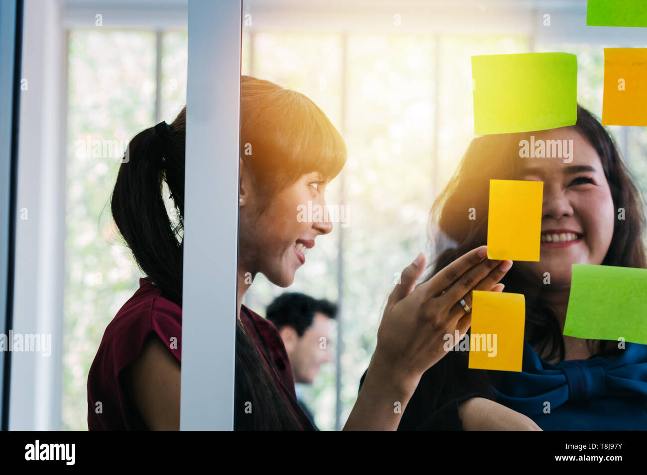 Female office managers smiling while looking at note stickers on glass wall in modern office