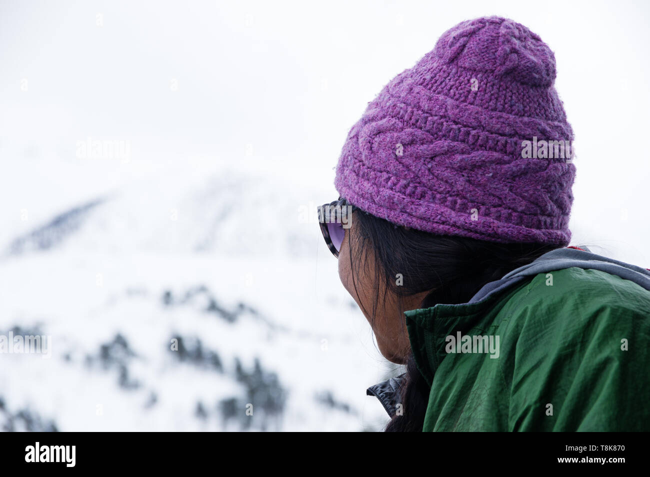 Asian woman in purple hat looks up towards out of focus snowy mountain on overcast winter day Stock Photo