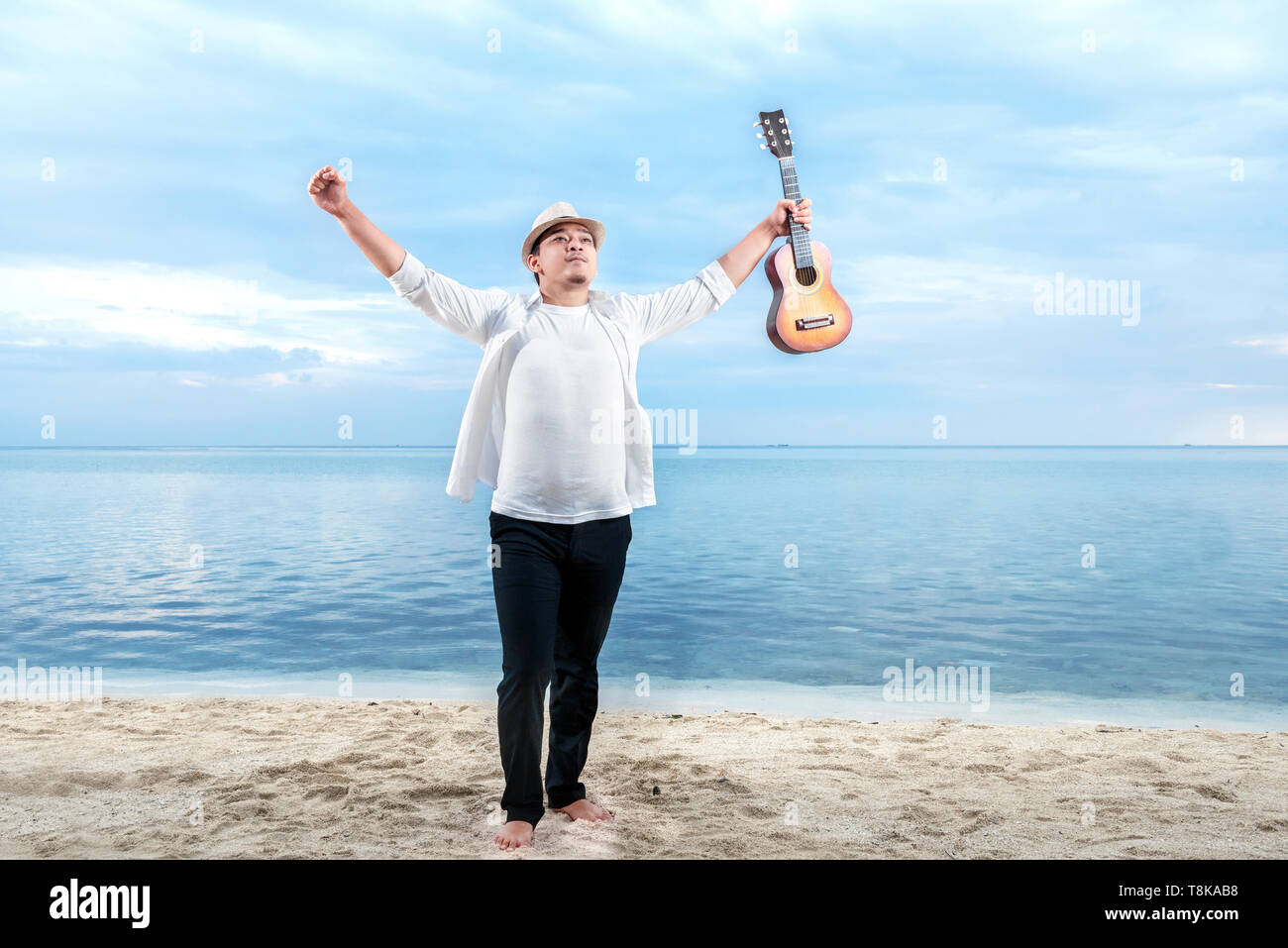 Asian businessman in hat and white clothes raised both hands while holding guitar on the beach. Summer vacation Stock Photo