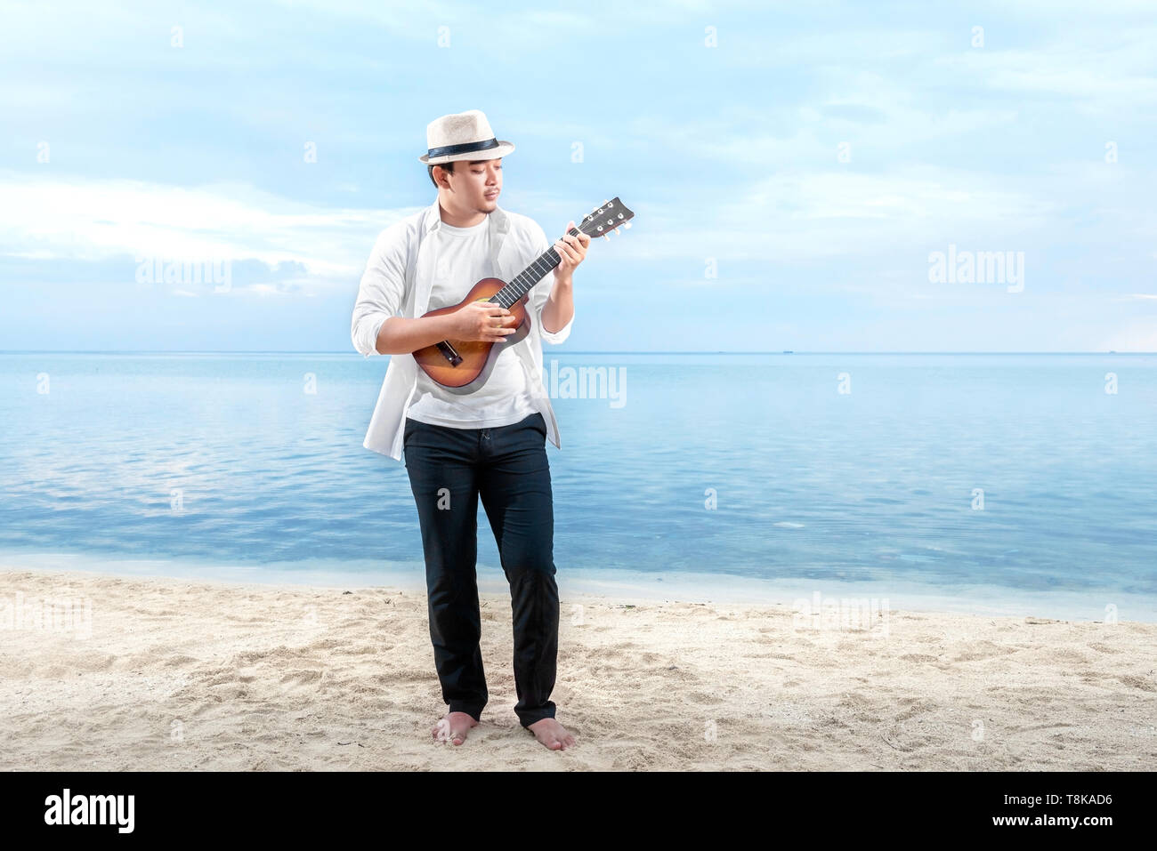 Asian businessman in hat and white clothes playing guitar on the beach. Summer vacation Stock Photo