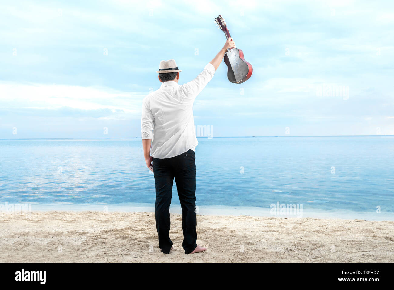 Rear view of asian businessman in hat and white clothes holding guitar on the beach. Summer vacation Stock Photo