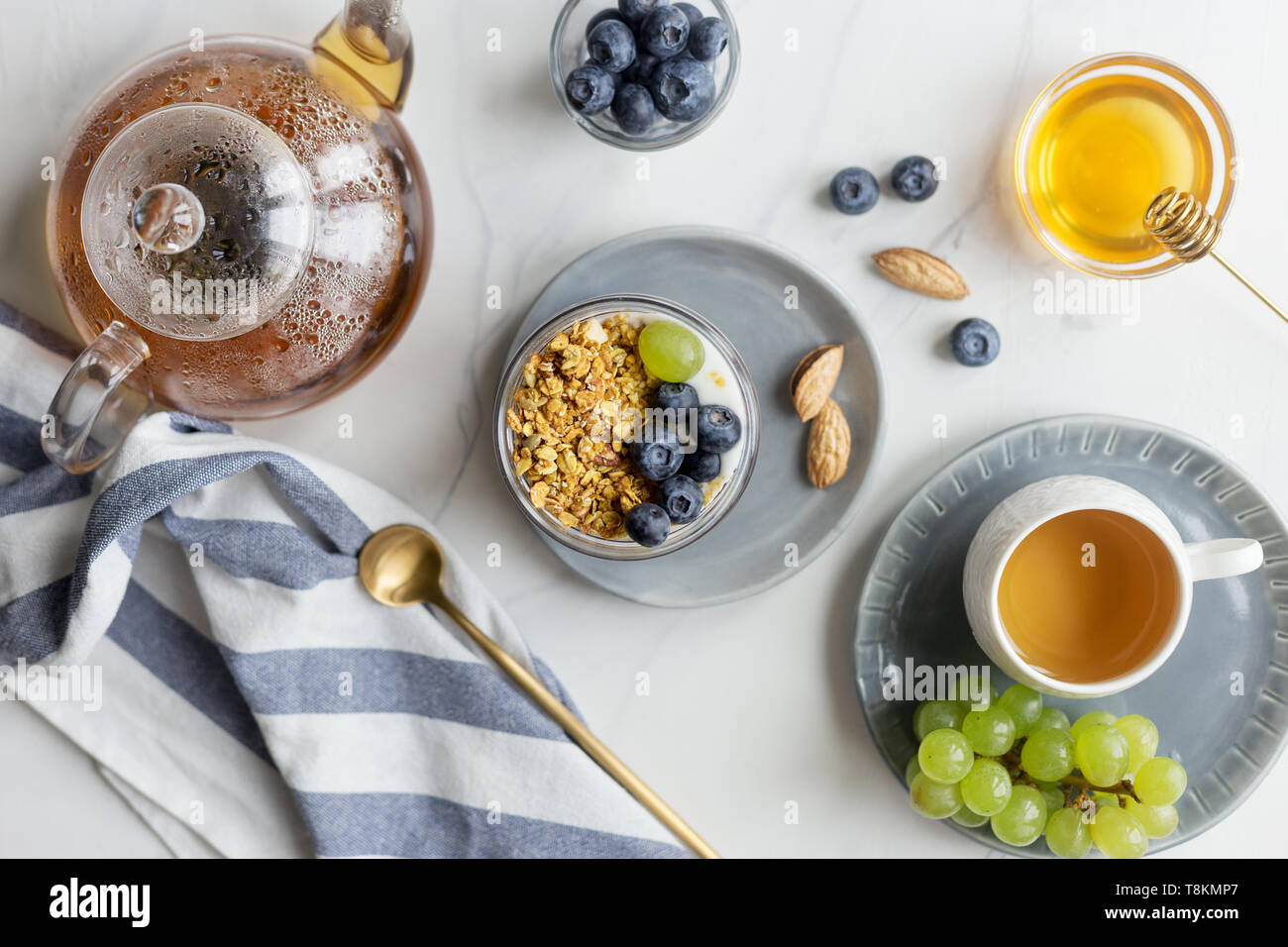 Summer tasty breakfast with granola, blueberry and grape with milk, honey and teacup on white marble background with napkin. Concept of healthy breakf Stock Photo