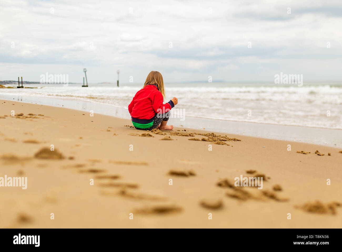 Young child sat on beachs waters edge lost in thought, taken from behind. Poole, Dorset, England. Stock Photo