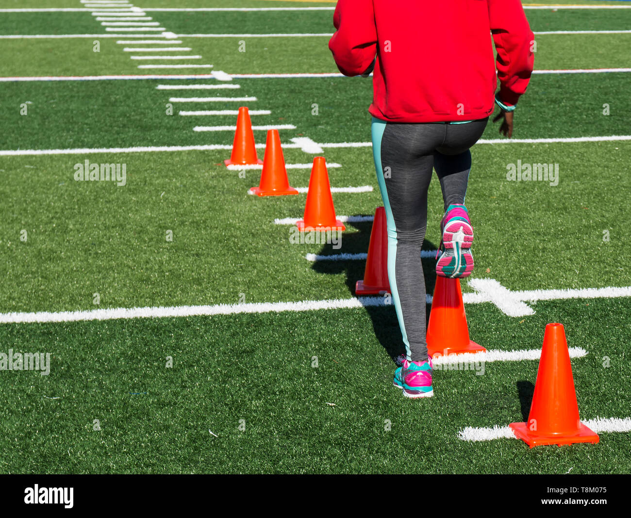A high school female athlete performs running drills over orange cones on a green turf field on a bright sunny fall afternoon wearing gray spandex. Stock Photo
