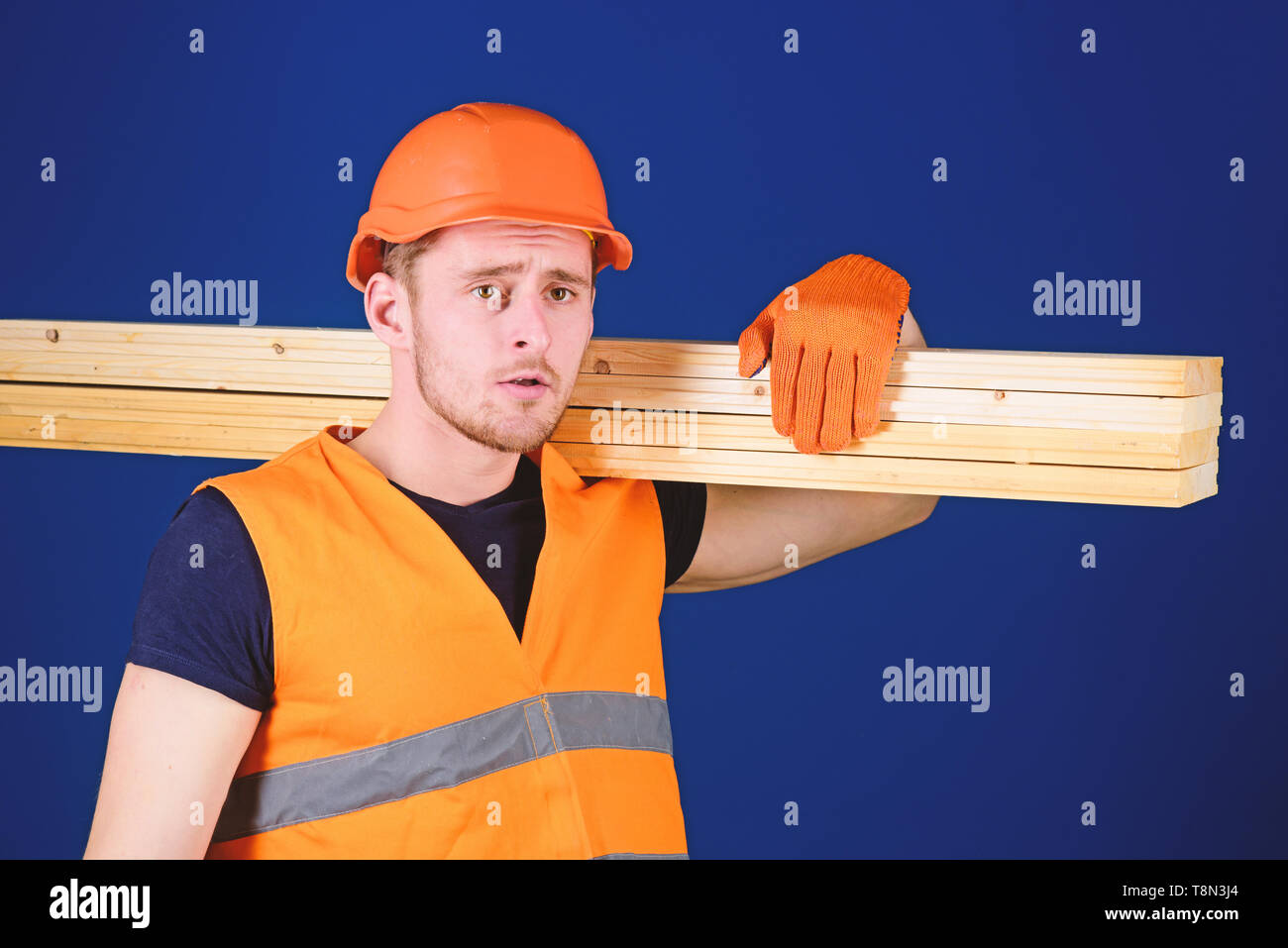 Man in helmet, hard hat and protective gloves holds wooden beam, blue background. Wooden