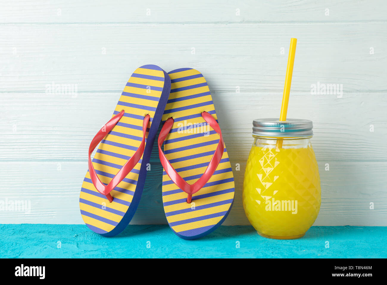 Flip flops and glass jar with fresh juice on color table against white wooden background, space for text. Summer vacation backdrop Stock Photo