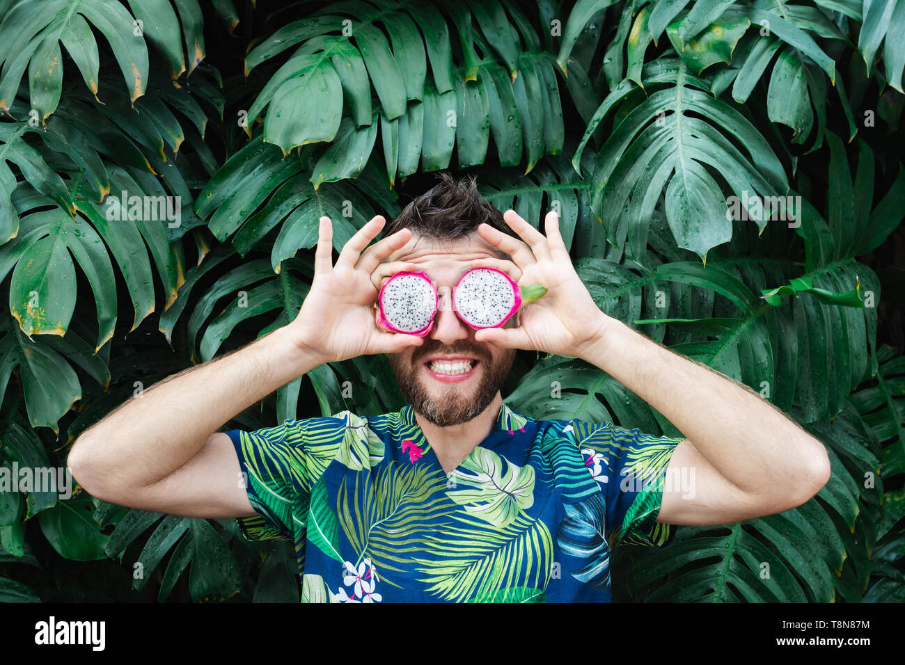 Young bearded man holding slices of Pitaya dragon fruit in front of his eyes, laughing. Tropical leaves background, copy space. Stock Photo