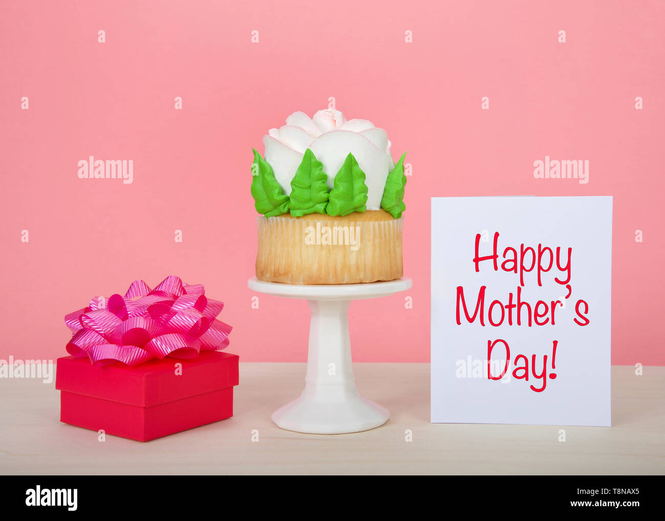 Large cup cake with giant frosting rose sitting on white pedestal on wood table with bright present and card with Happy Mother's Day card on the table Stock Photo