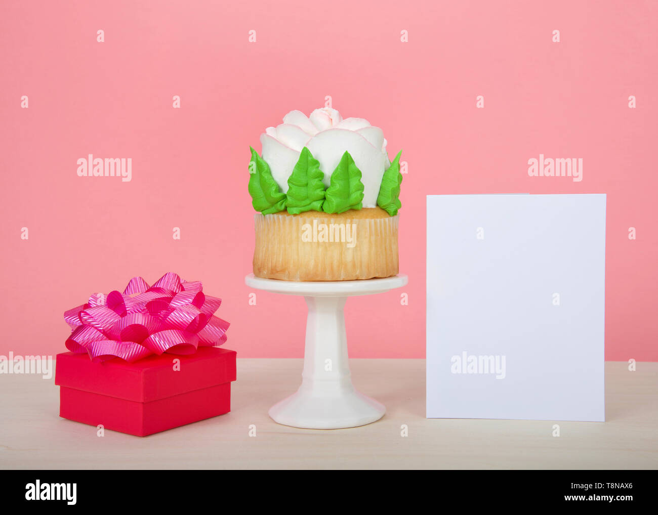 Large cup cake with giant frosting rose sitting on white pedestal on wood table with bright present and blank card, copy space Stock Photo