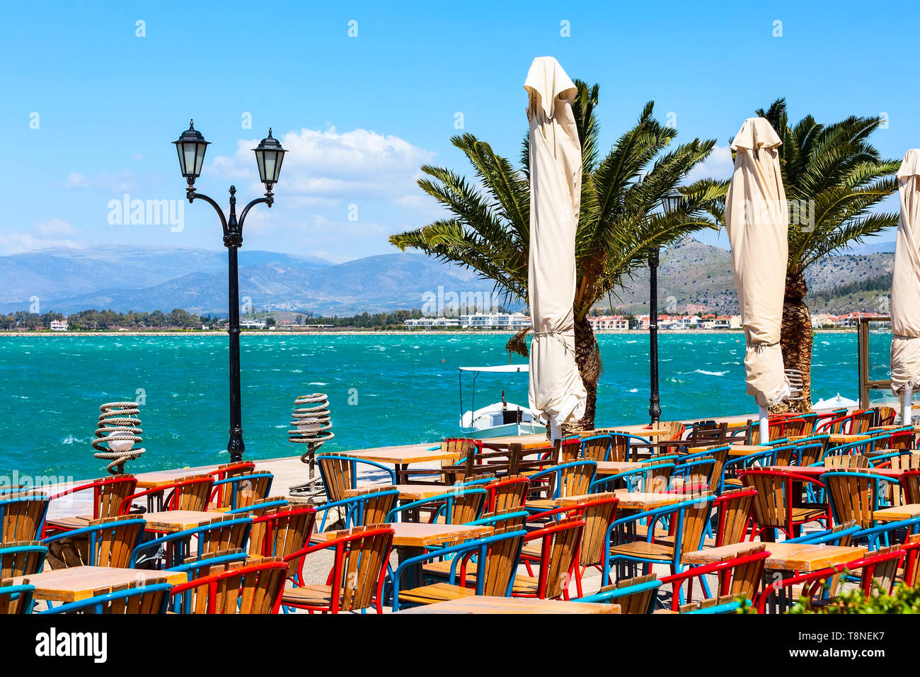 Promenade with colorful tables and chairs in street tavern restaurant, palm trees and sea in Nafplio or Nafplion, Greece, Peloponnese Stock Photo