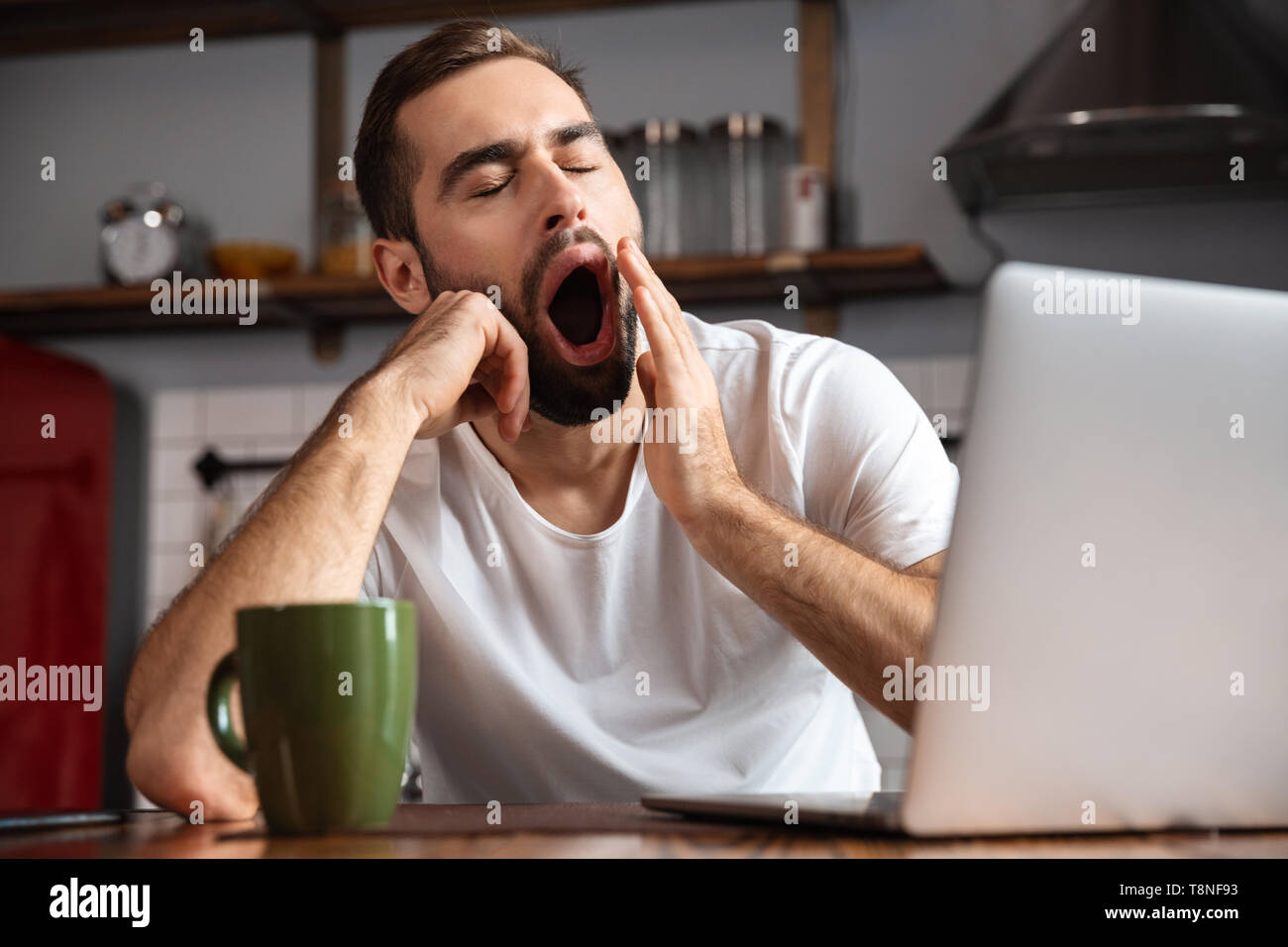 Bored young man using laptop computer while sitting at the kitchen table Stock Photo