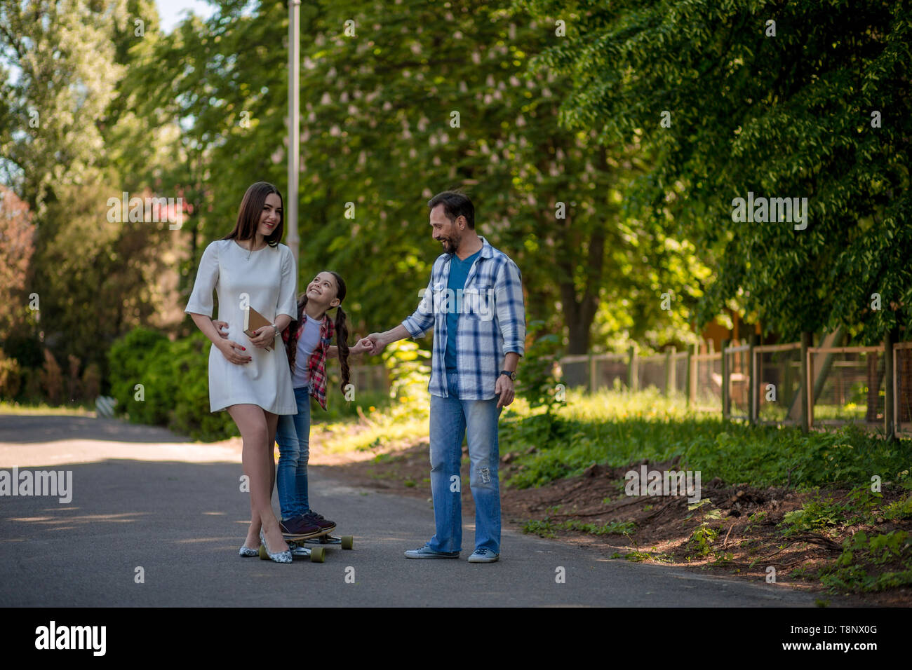 Family Spend Time In The City Park. Father Is Helping Daughter To Ride On The Skateboard In The Park. Stock Photo