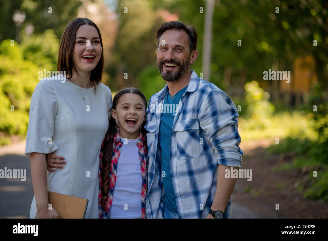 Smiling Happy Family In The Park. Mother Father And Daughter In Casual Clothes. The Daughter Is Hugging Her Parents. Stock Photo