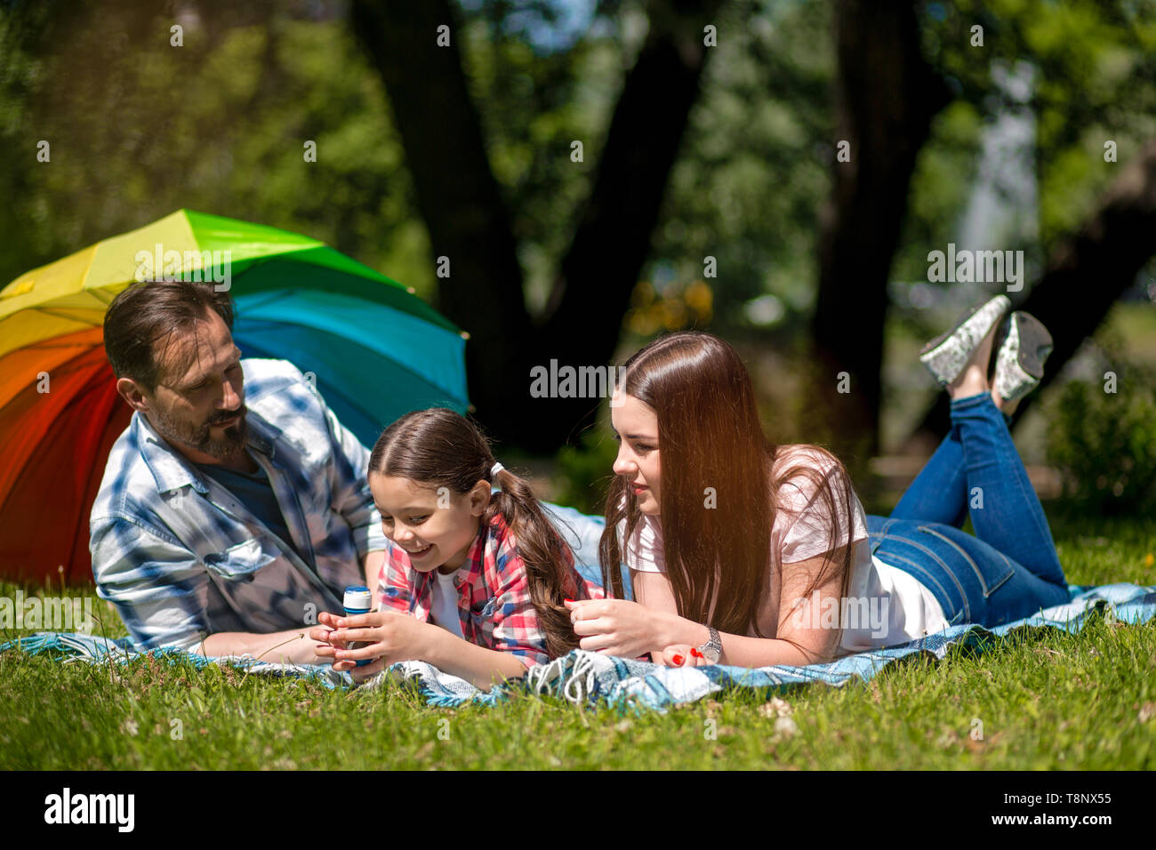 Family Lying Together On A Picnic Blanket Outdoors In The Park. Colorful Umbrella On The Background. Stock Photo