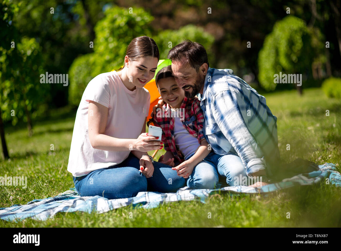 Young Woman Is Showing Photos On Phone To Her Husband And Daughter While Sitting On The Blanket In The Park. Stock Photo