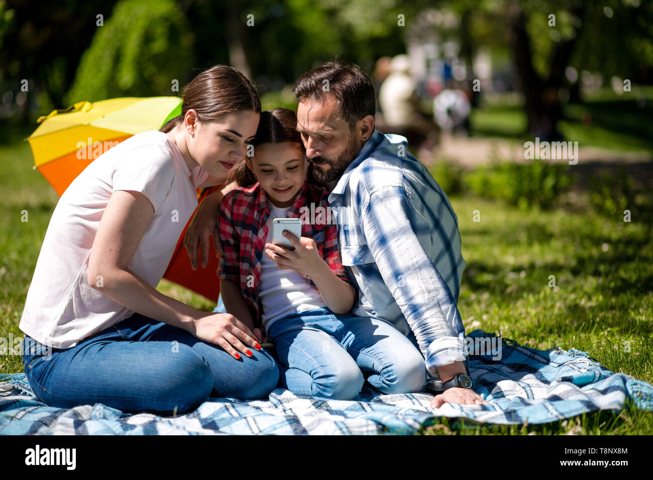 Smiling Family Watching Photo On Mobile Phone While Sitting On The Blanket In The Park. Stock Photo