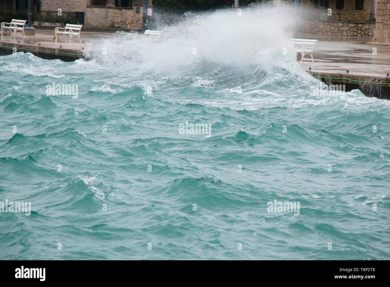 Stormy sea splashing the waves on coast promenade with benches in Dalmatia seaside town in off season during strong south wind with rain Stock Photo
