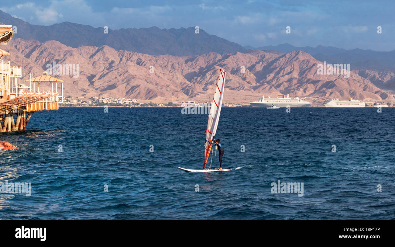 a child windsurfer sailing into the eilat marina in israel with the port of akaba jordan in the background Stock Photo
