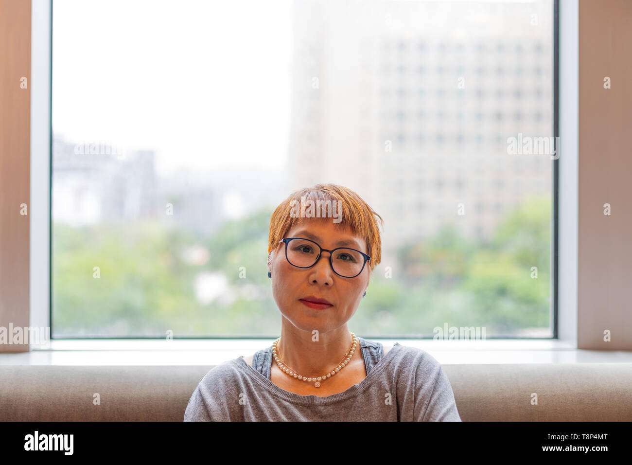 Mature Taiwanese woman of Chinese ethnicity sitting in front of a window looking into the camera Stock Photo
