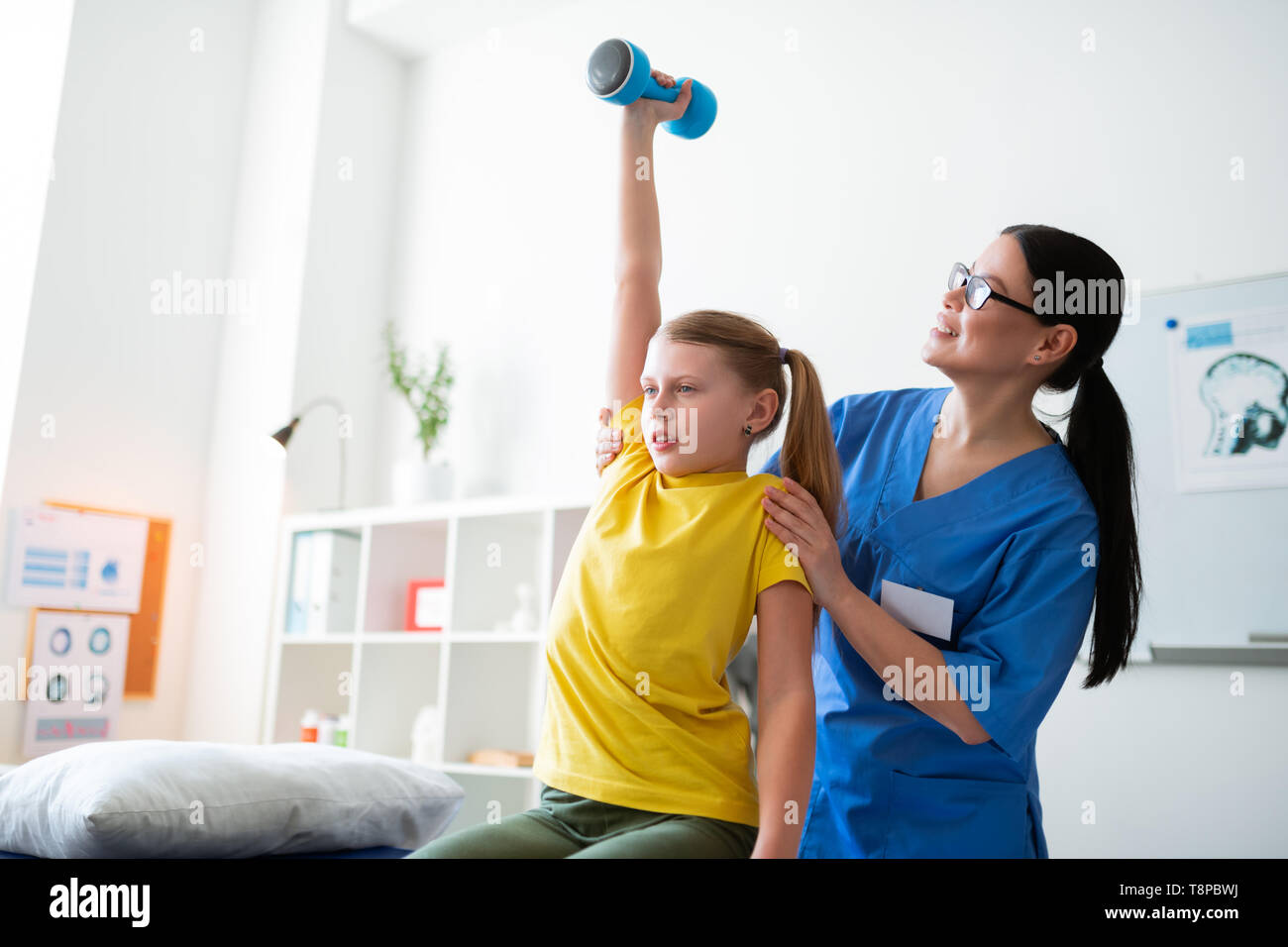 Concentrated skinny girl sitting on the daybed and raising dumbbells Stock Photo