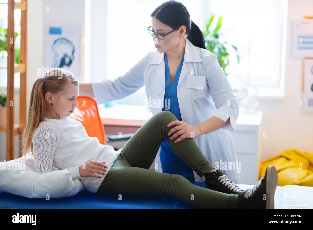 Blonde kid lying on a daybed while doctor observing her hurting leg Stock Photo